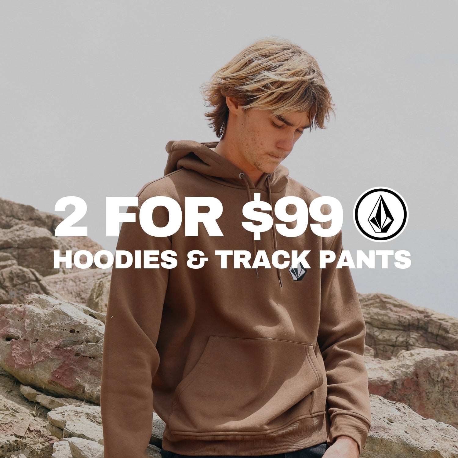 A man wearing a brown Volcom hoodie stands outdoors against a rocky, overcast sky. Large white text across the center reads '2 FOR $99 HOODIES & TRACK PANTS' with the Volcom logo.