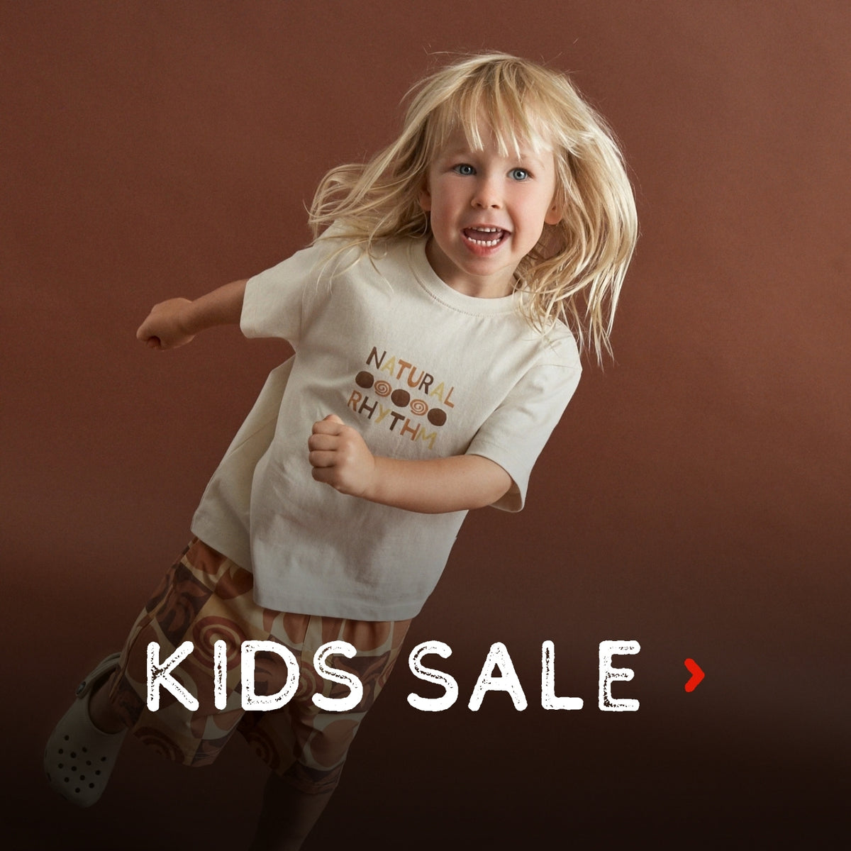 A young blonde child wearing a cream graphic t-shirt and patterned shorts jumps against a solid dark brown background. The t-shirt features the text 'NATURAL RHYTHM' in brown and gold.