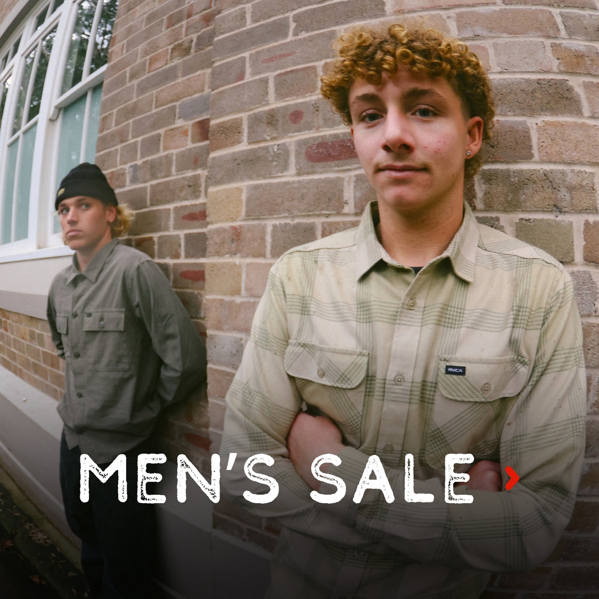 Two young men standing against a brick wall outdoors, with the text 'MEN'S SALE' overlaid across the bottom. The man in the foreground wears a light beige plaid button-up shirt, and the man in the background wears a dark gray shirt and black beanie.