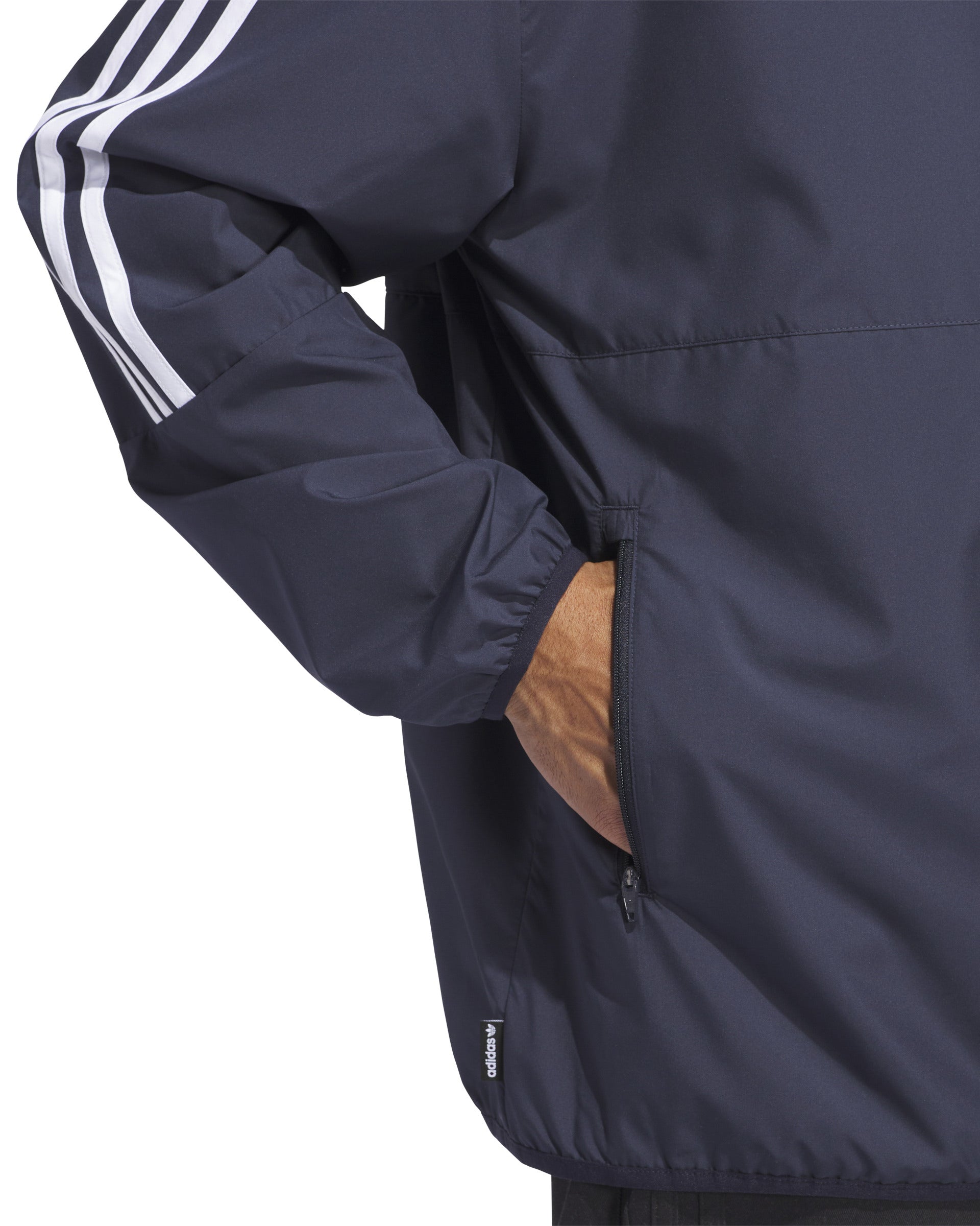 Close-up of a navy Adidas windbreaker jacket with white stripes on the sleeve, showing a hand in the zippered side pocket.