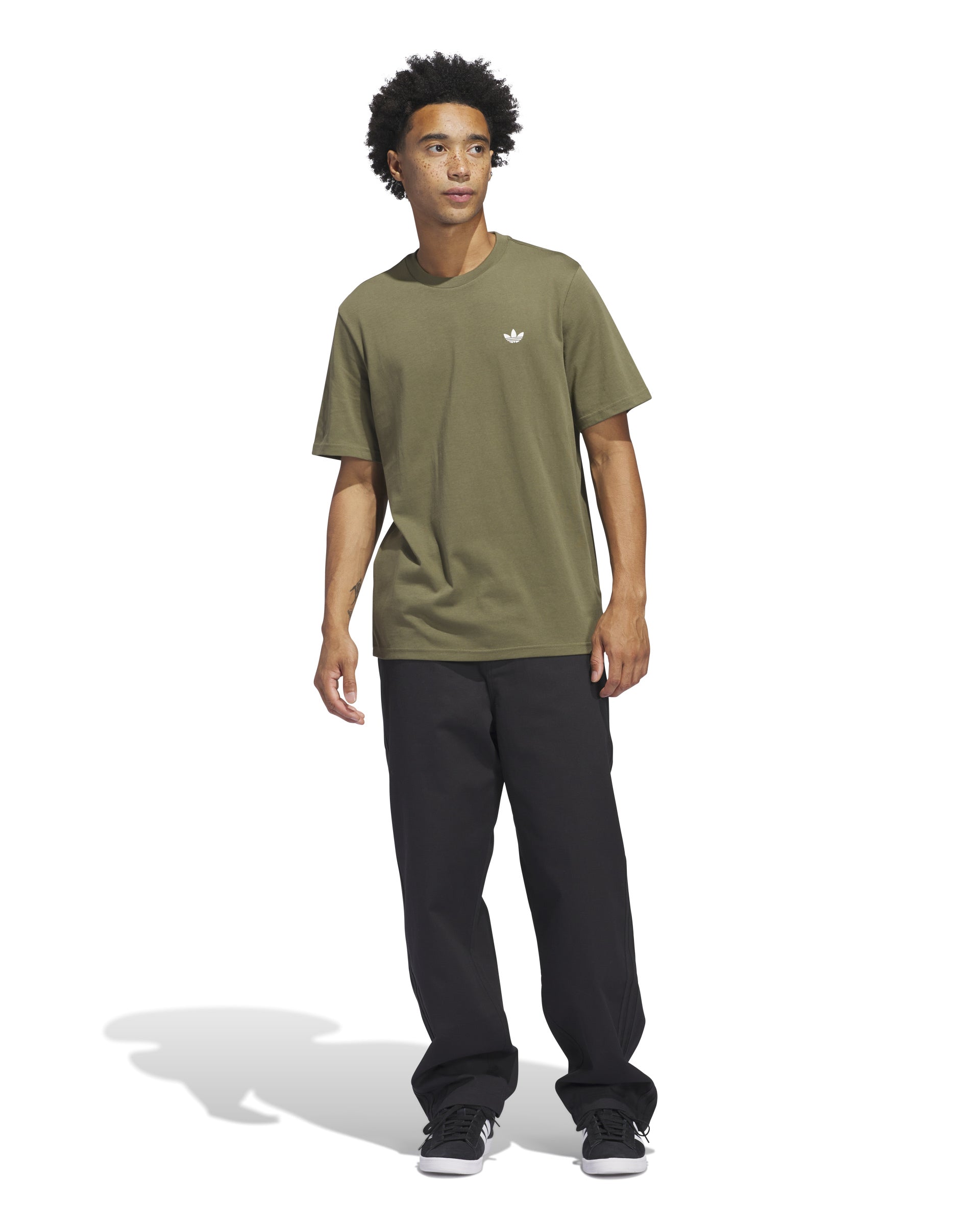 Full-body view of a model wearing an olive green Adidas short sleeve tee with a white logo, black pants, and black sneakers.