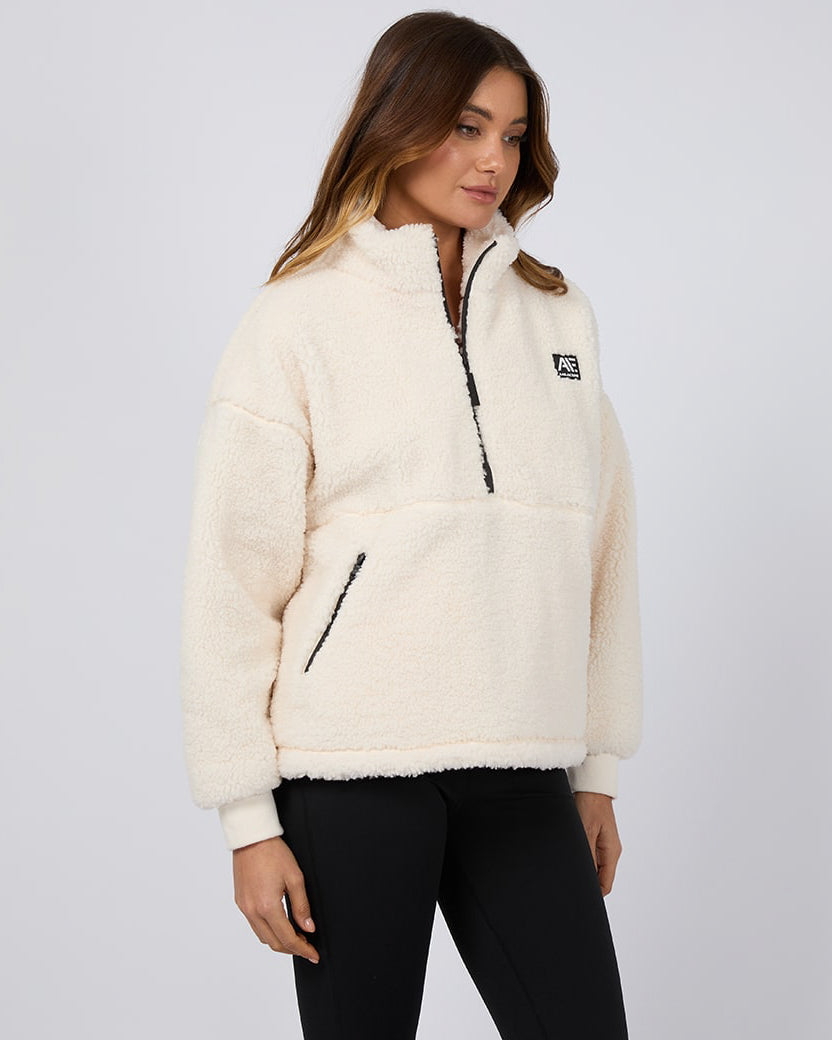 Side view of a cream sherpa fleece pullover with a half-zip front, black logo patch, and zippered pocket, worn by a model.