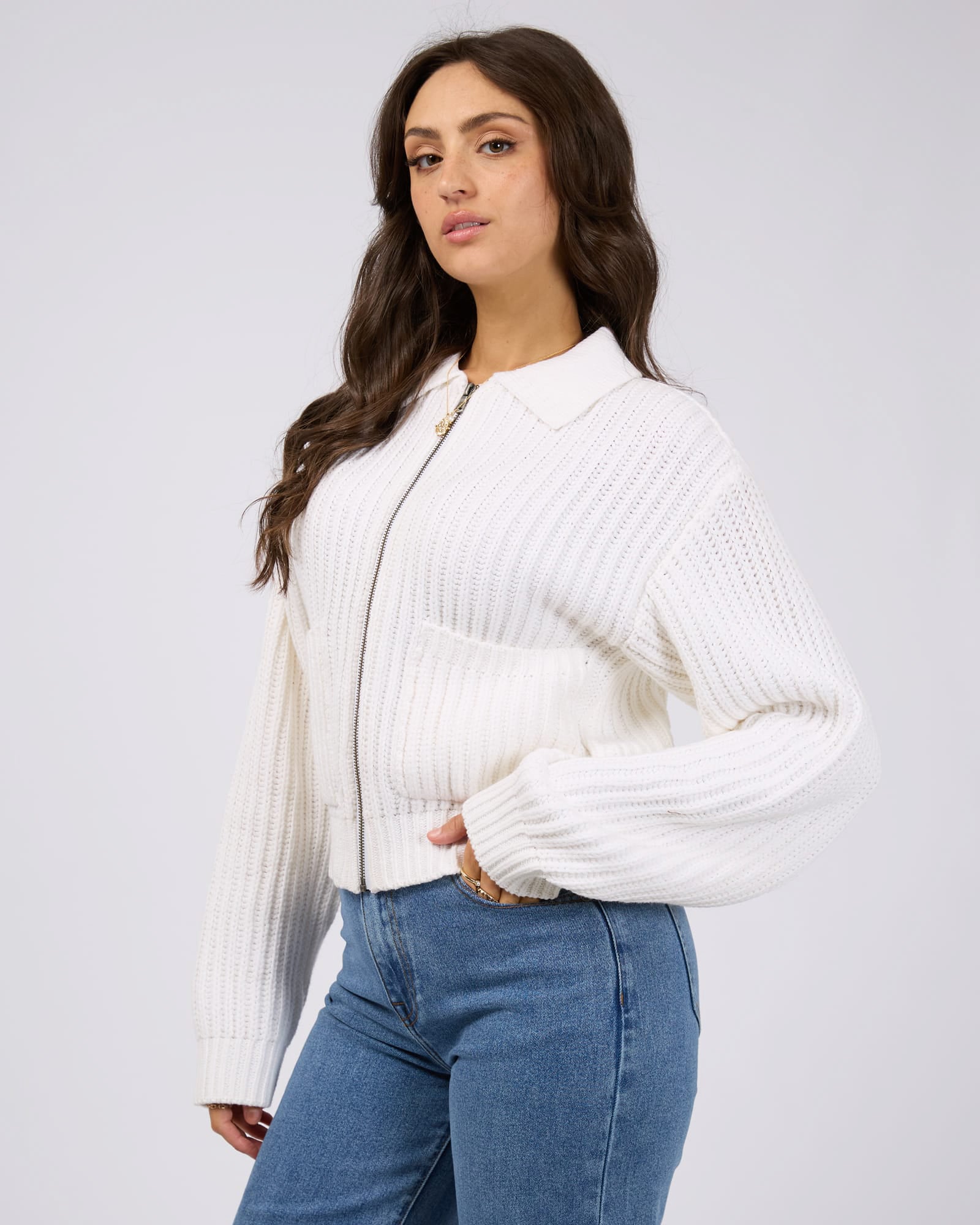 Side view of a model wearing a white ribbed knit zip-up cardigan with a collar and balloon sleeves, paired with blue jeans.