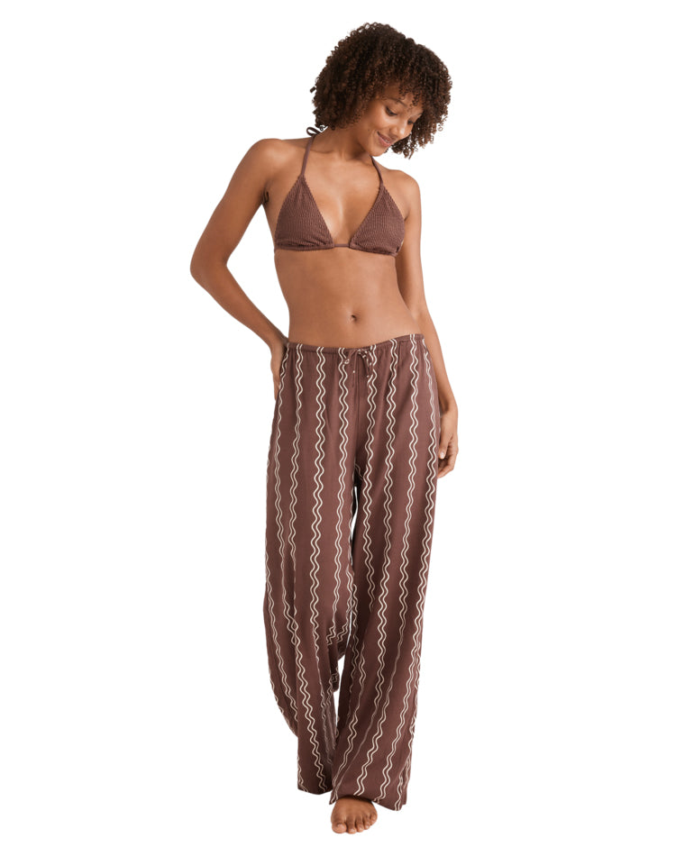 Woman in brown zigzag print pants and brown bikini top, looking down with one hand in pocket, barefoot.