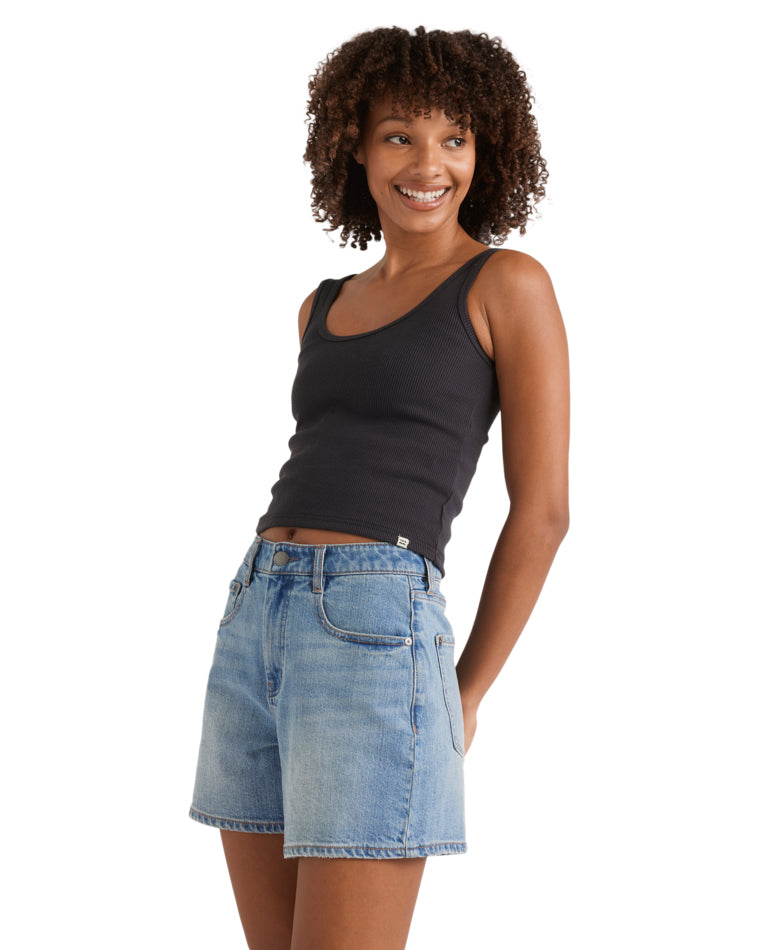 Side view of woman in black ribbed tank and high-waisted mid-blue denim shorts with hands in pockets.