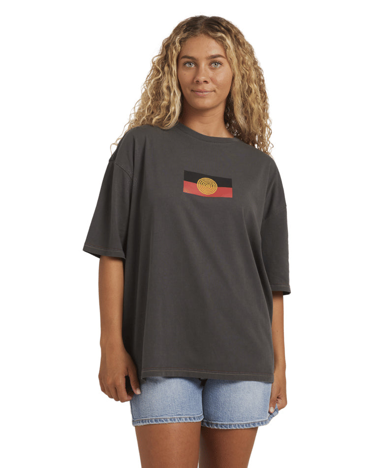 Model wearing a black Billabong t-shirt featuring an Aboriginal flag graphic on the chest, paired with denim shorts.