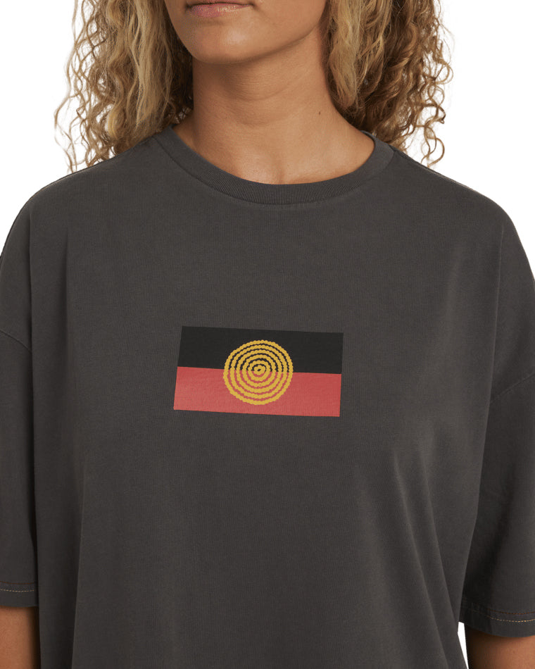 Close-up of Aboriginal flag graphic on the chest of the black Billabong t-shirt, featuring concentric yellow circles.