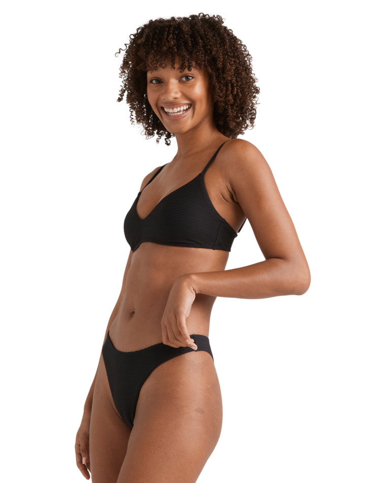 Side view of a woman in a black bikini smiling, showing adjustable bra-style top and high-leg bikini bottoms.