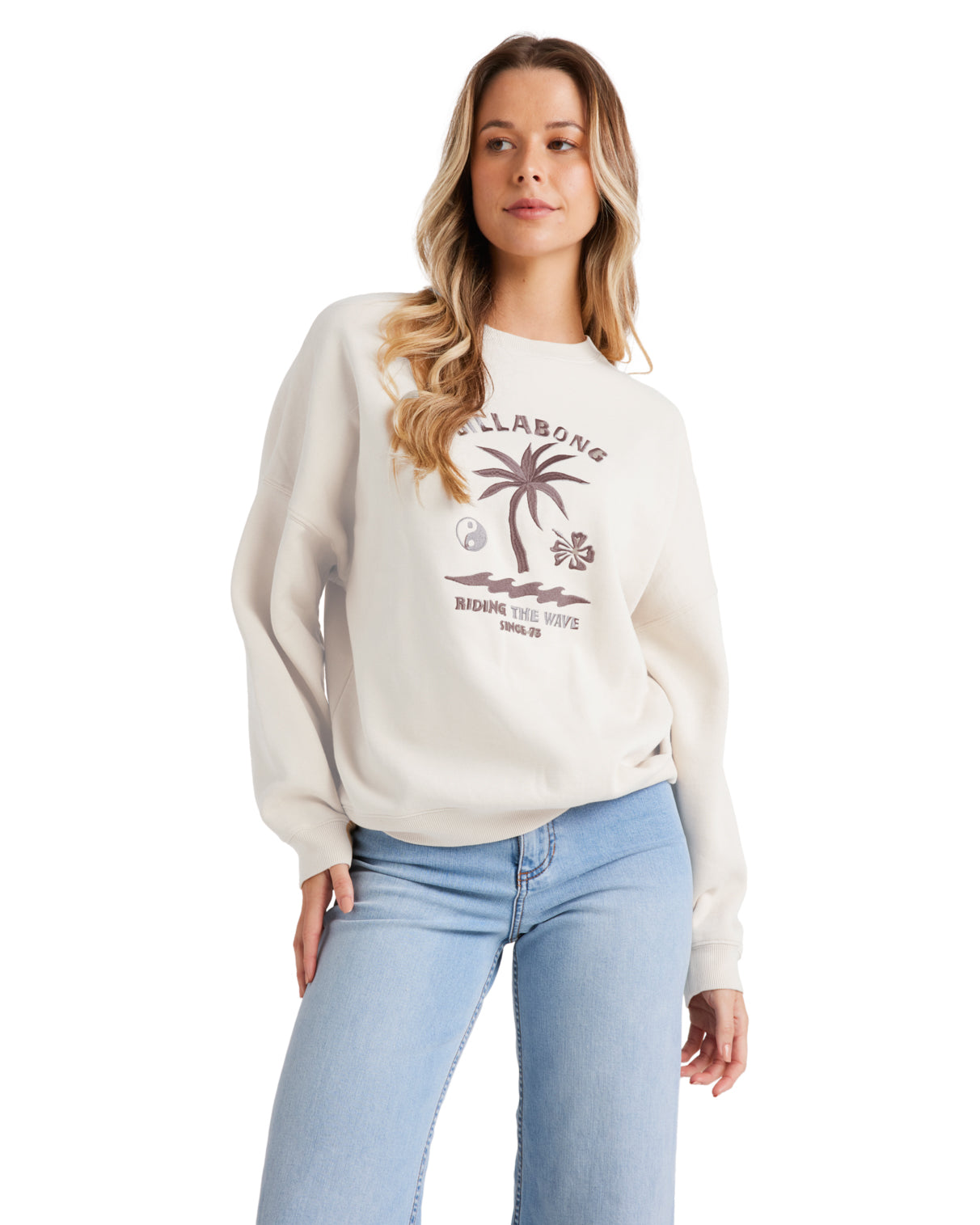 Model wearing a cream Billabong sweatshirt with a tropical palm tree graphic, paired with light blue jeans.