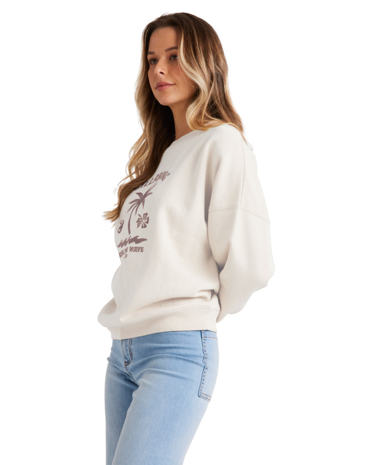 Model wearing a cream Billabong sweatshirt with a tropical graphic, paired with light blue jeans, shown from the side.