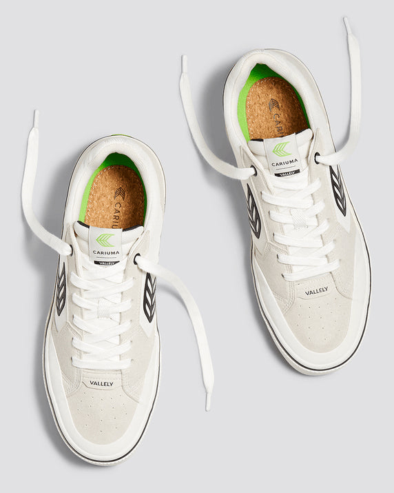 Top view of white Cariuma Vallely sneakers with black details, cork insoles, green branding, and untied laces on a grey background.