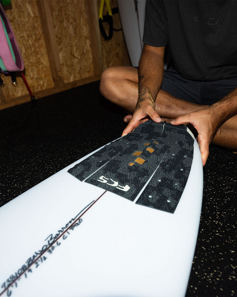 Surfer applying black camo FCS tail pad to white board in a surf shed, highlighting pad shape and traction grip.