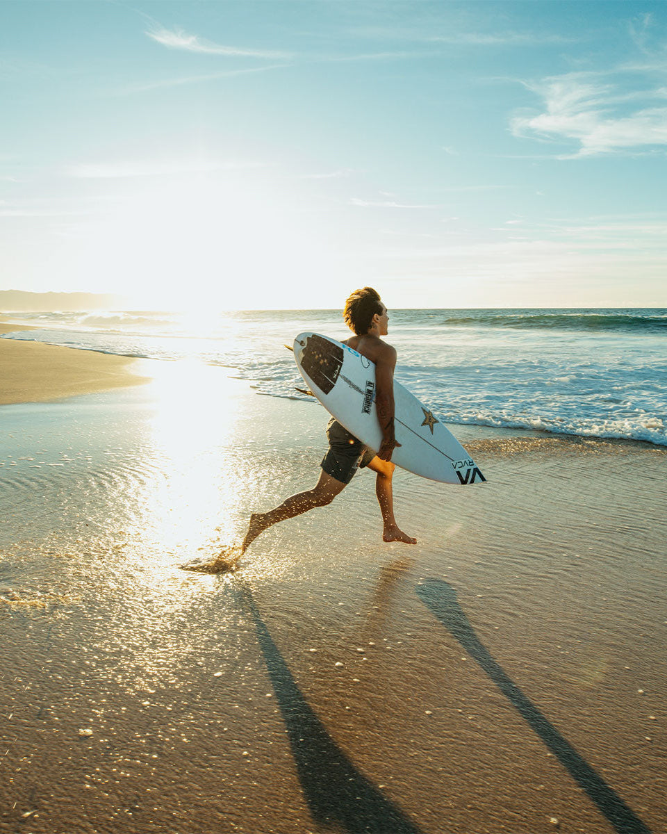 Surfer running along shoreline with surfboard featuring black camo FCS tail pad under sunset light.