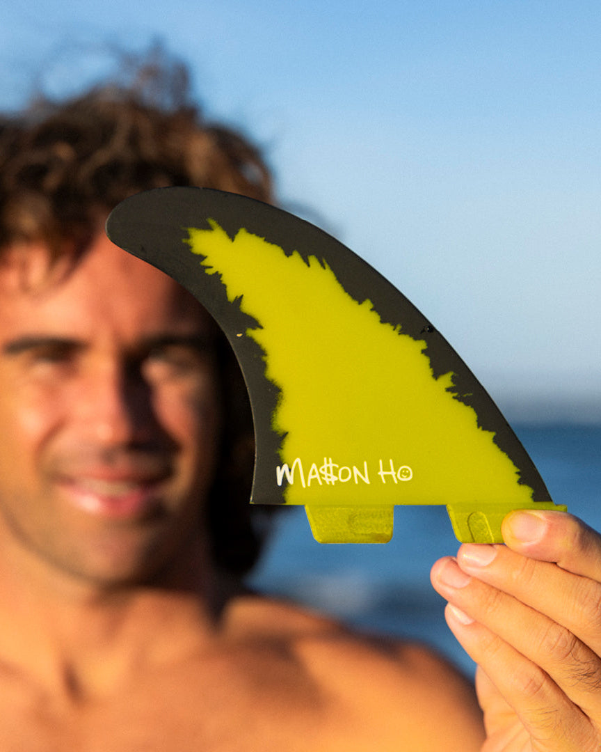Surfer holding up FCS II Mason Ho fin, showcasing its signature black and yellow design near the beach.