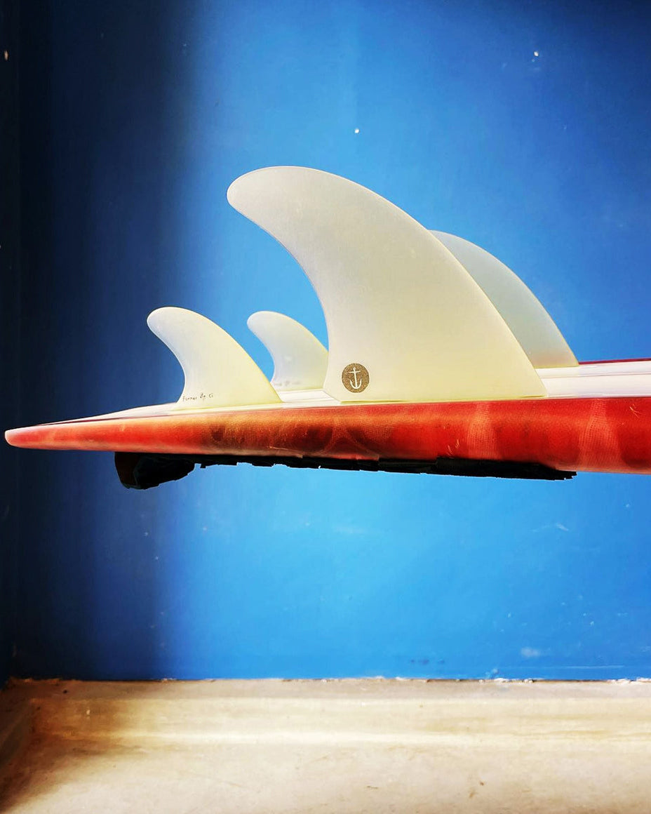 Side view of a red Channel Islands surfboard highlighting translucent quad fins with anchor emblem and slight rocker.