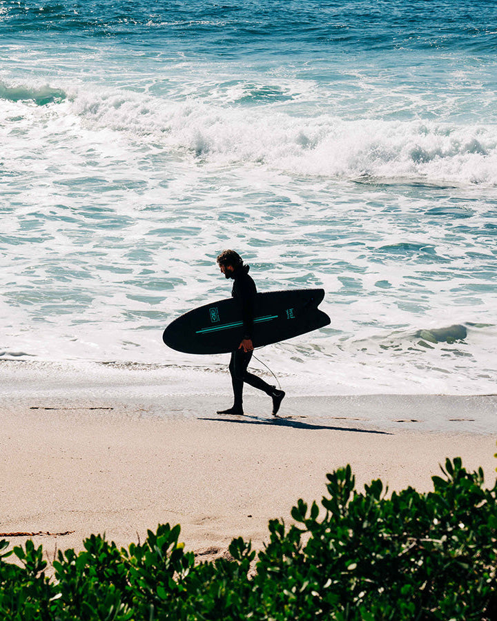 Surfer walking along beach carrying navy Ocean & Earth Flying Fish board.