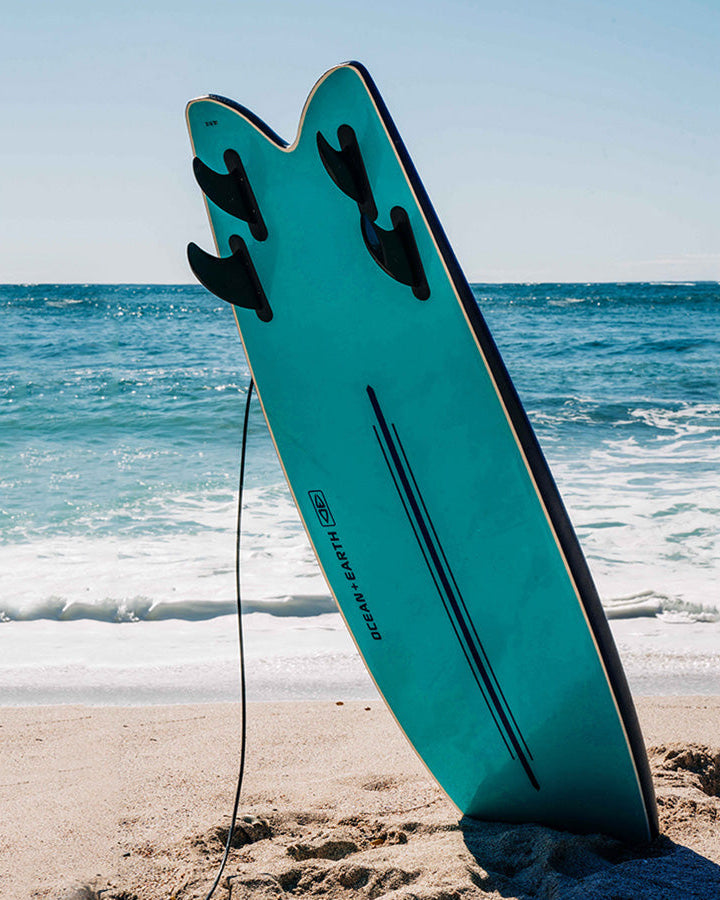 Ocean & Earth softboard with fins standing upright on sandy beach.