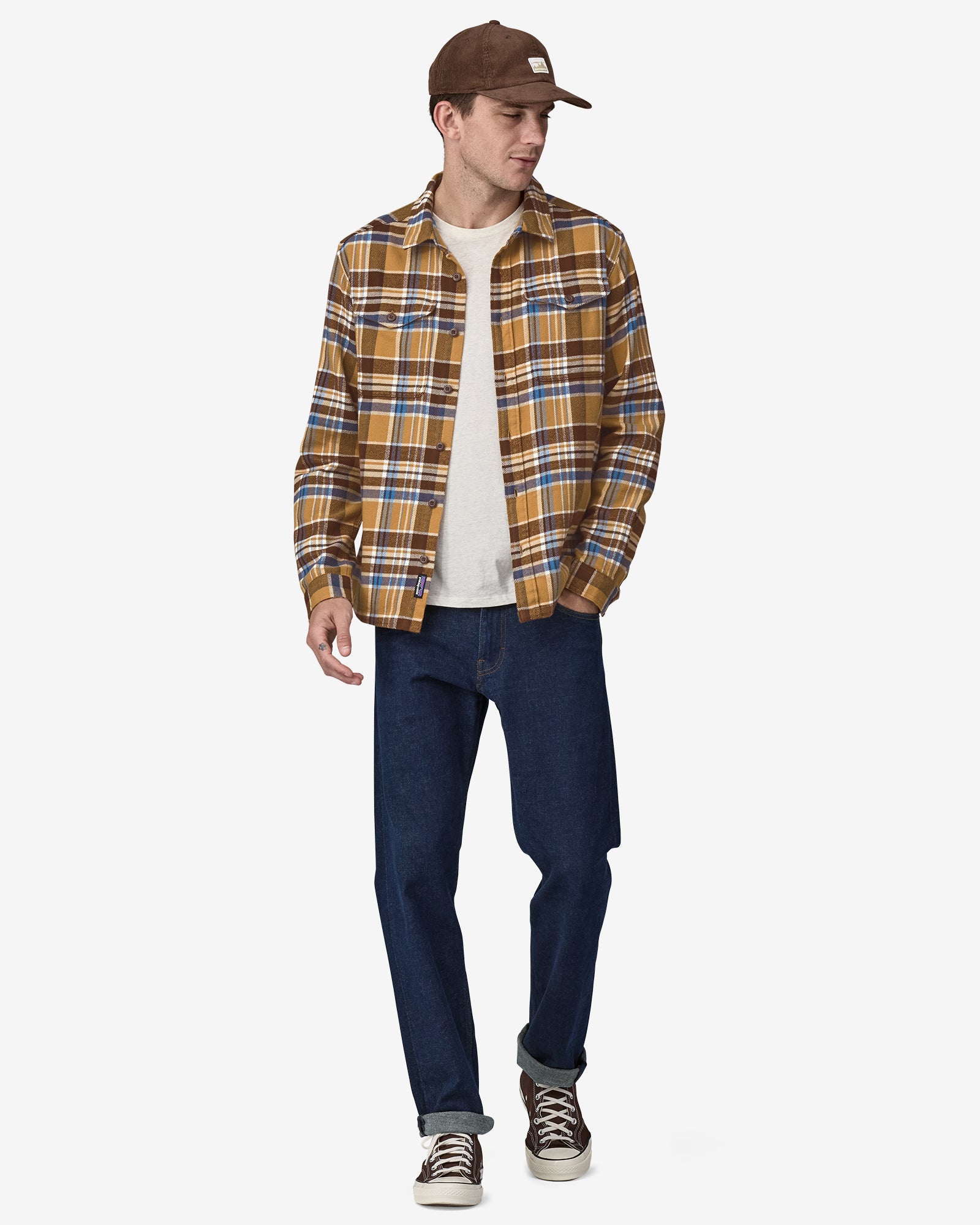 Man wearing a mustard, brown, and blue plaid flannel shirt over a white tee, paired with dark jeans, sneakers, and a cap.