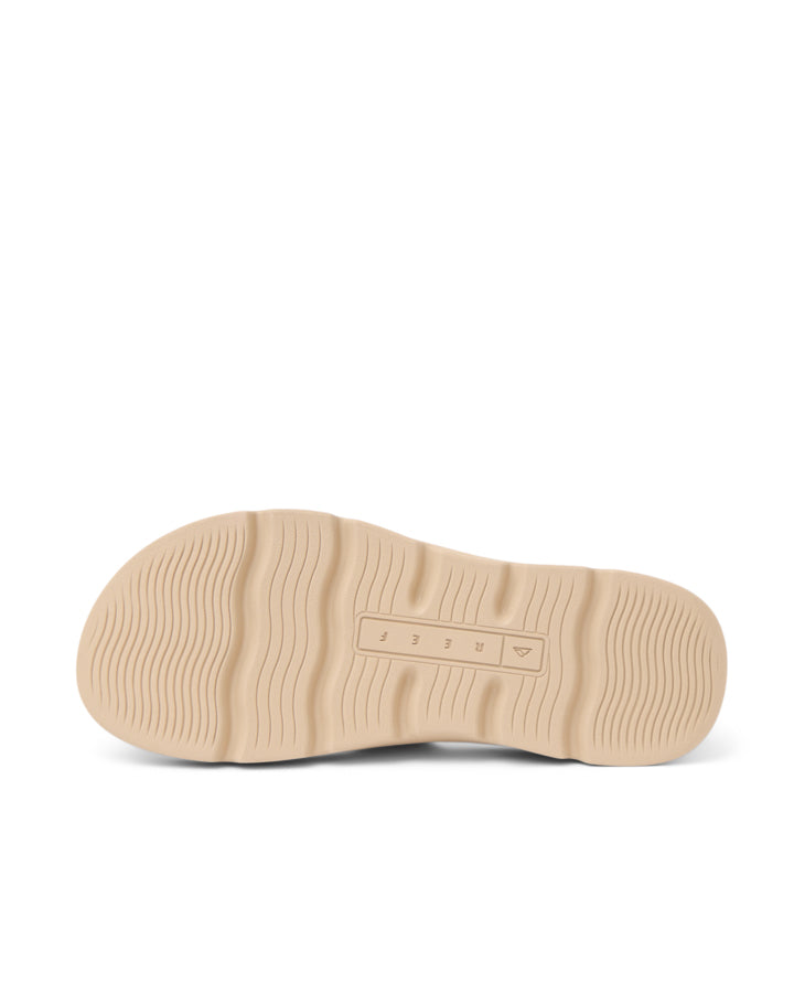 Underside view of a beige slide sandal showing grooved sole and embossed Reef logo.