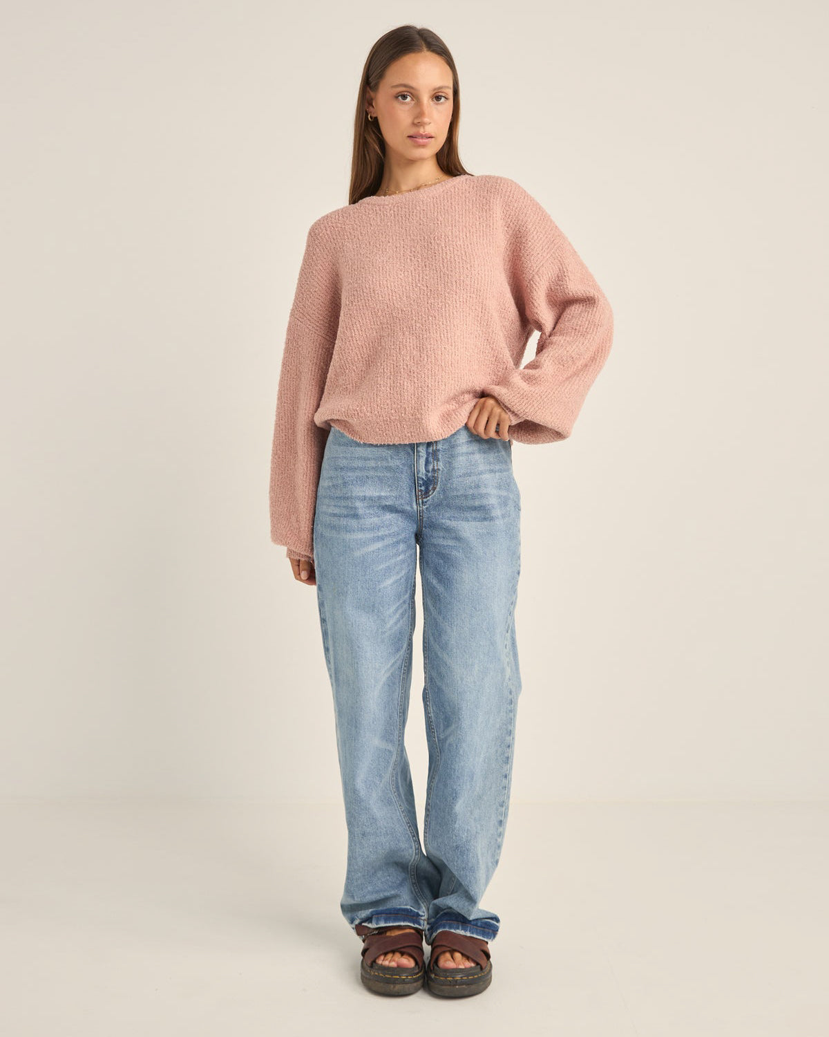 Woman wearing a pink oversized knit sweater, light blue relaxed-fit jeans, and brown sandals, standing against a neutral background.