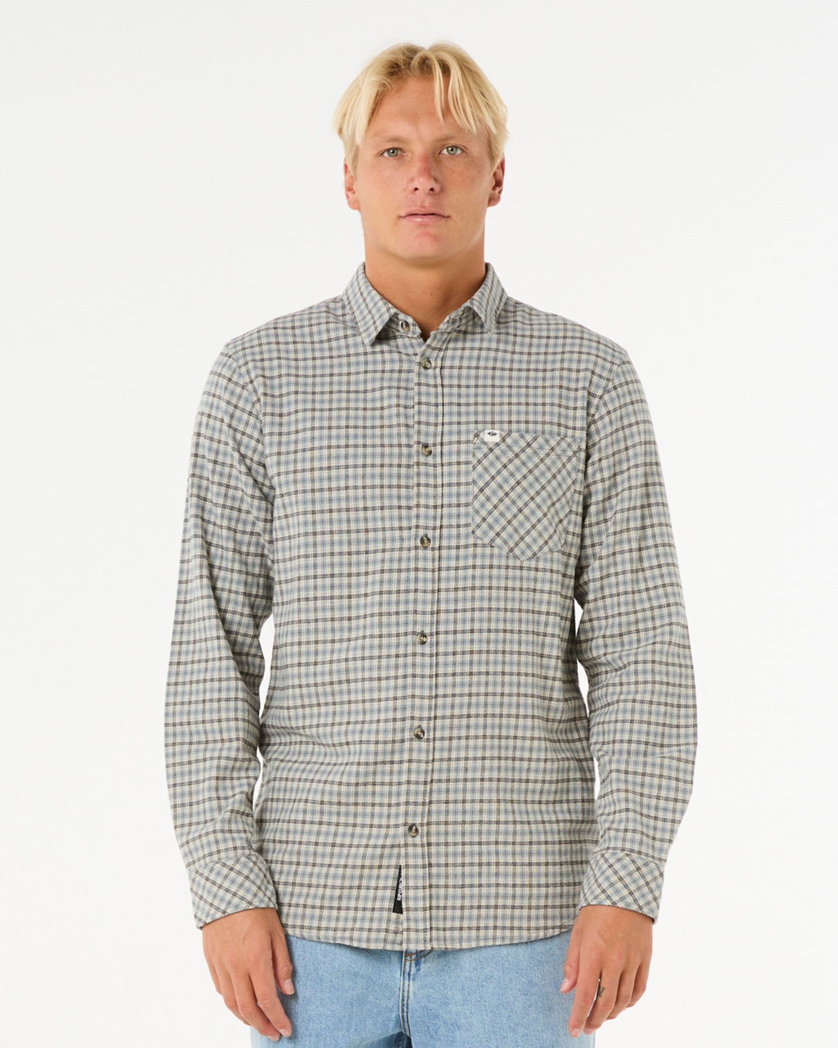 Man wearing a long-sleeve button-up plaid shirt in neutral tones with a front chest pocket, paired with light blue jeans.