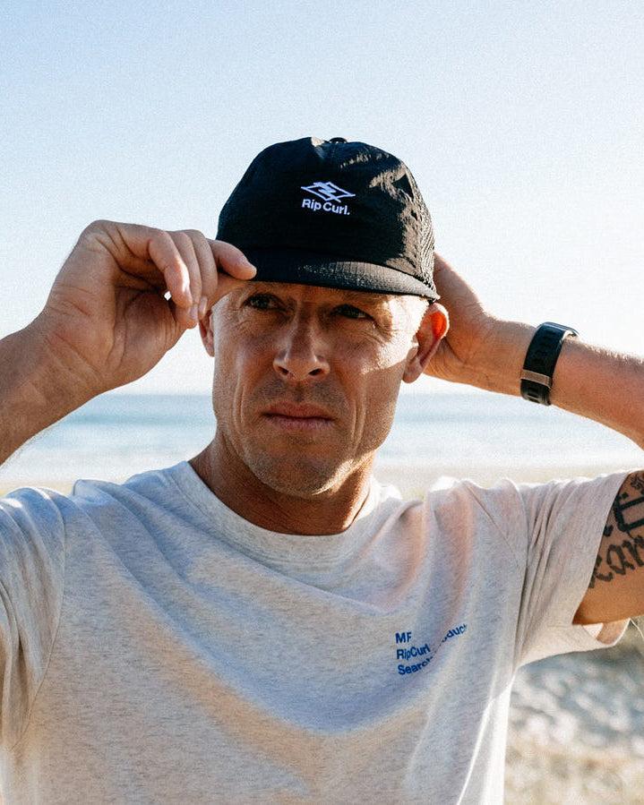 Man wearing a black Rip Curl mesh-back cap at the beach, adjusting the brim while looking into the distance.