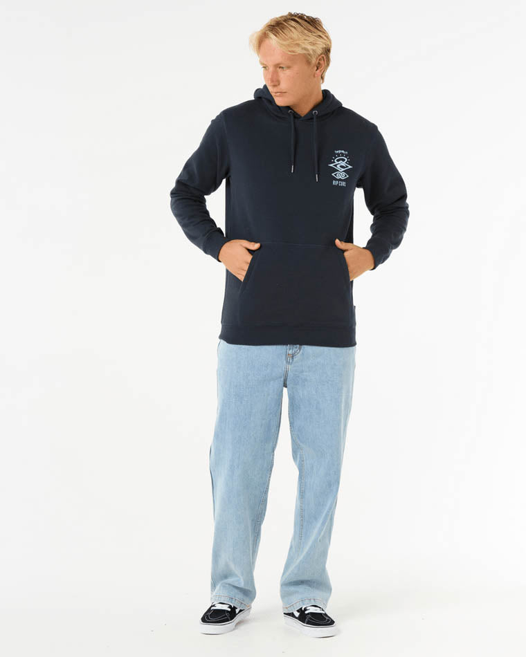 Man wearing a navy Rip Curl hoodie with a small white chest graphic, light blue jeans, and black sneakers.