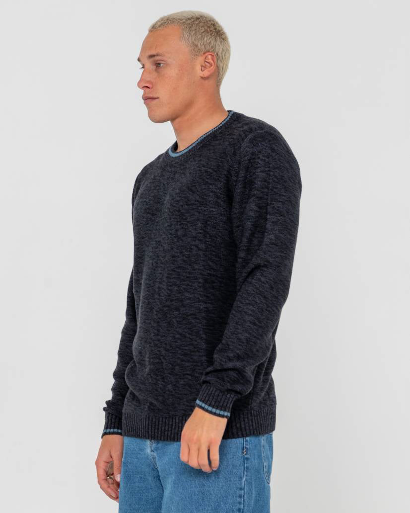 Side view of a male model wearing a dark navy knit sweater with ribbed cuffs and hem, paired with light blue denim jeans.