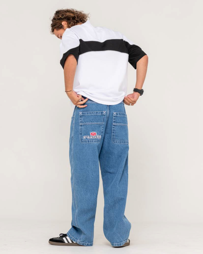 Rear view of model wearing loose-fit blue denim jeans with Rusty logo on back pocket, paired with a white and black t-shirt.