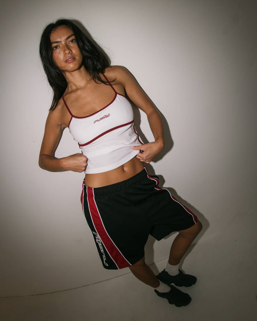 Female model wearing a white tank top with red piping and black athletic shorts with red accents, posing in a sporty outfit.