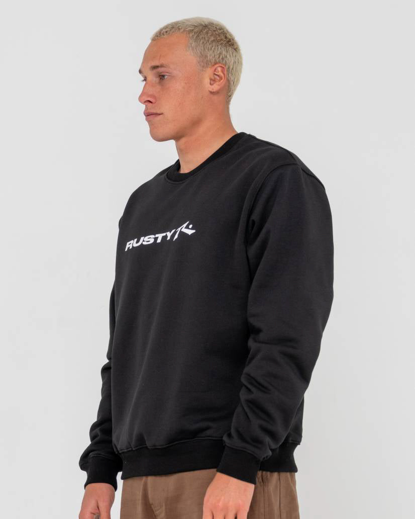 Side view of a man wearing a black Rusty crewneck sweatshirt with a white logo on the chest, paired with brown pants.
