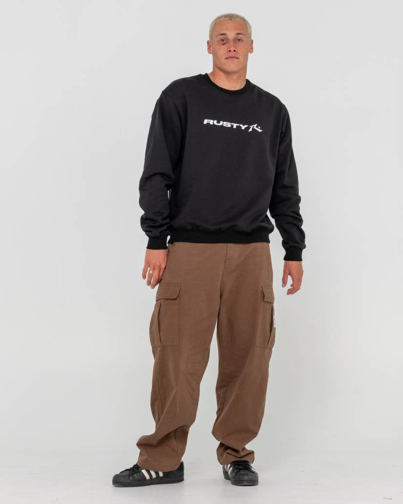 Man wearing a black Rusty crewneck sweatshirt with a white logo, brown cargo pants, and black sneakers, standing casually.