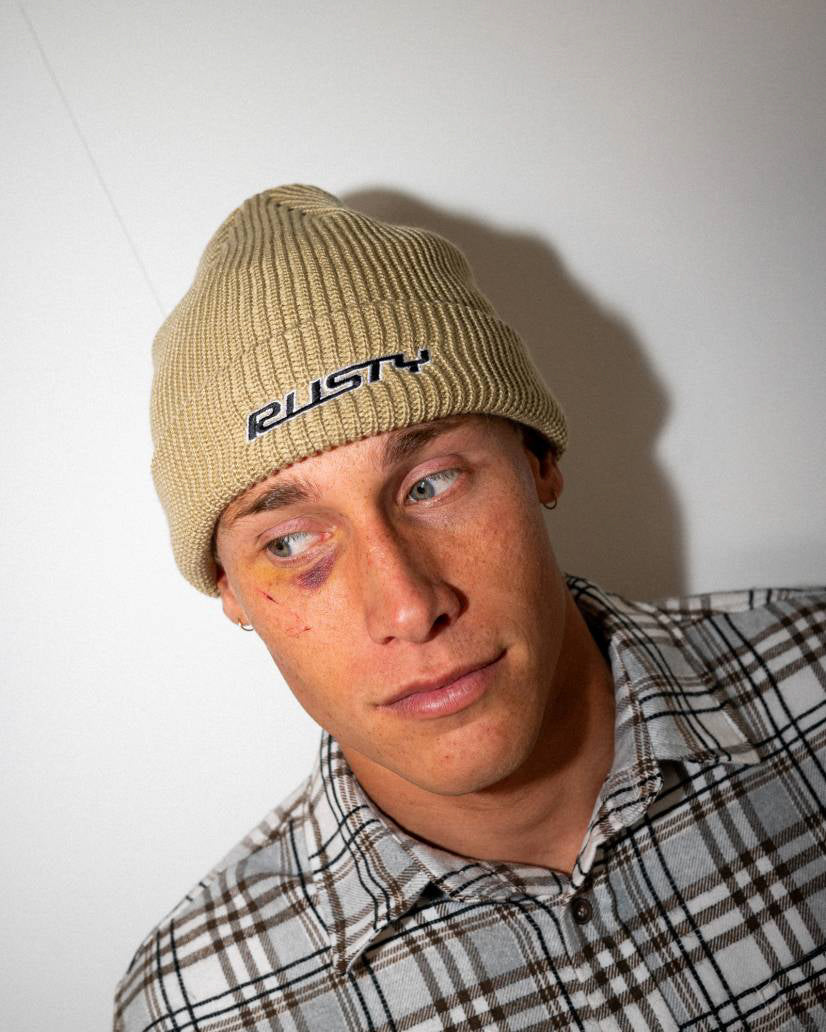 Man wearing a beige Rusty beanie and a plaid shirt, looking to the side with a neutral expression.