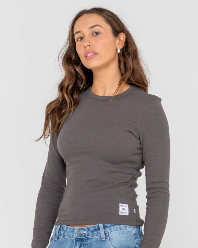 Model wearing a fitted, long-sleeve, ribbed dark grey top with a crew neckline and a small logo patch near the hem.