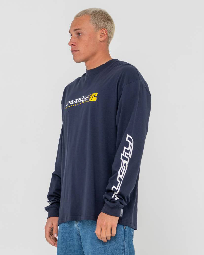Model wearing a navy blue Rusty long-sleeve shirt with a yellow logo on the chest and white branding on the sleeves, side view