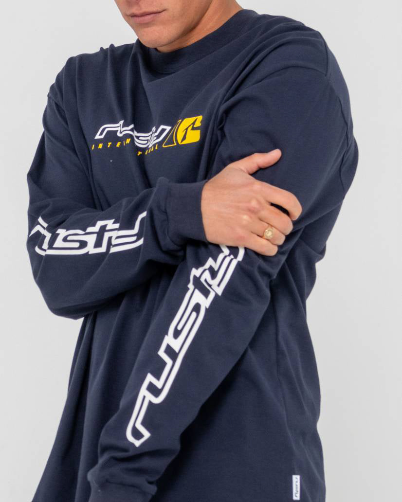 Close-up of a model wearing a navy blue Rusty long-sleeve shirt with a yellow chest logo and bold white branding on the sleeves.