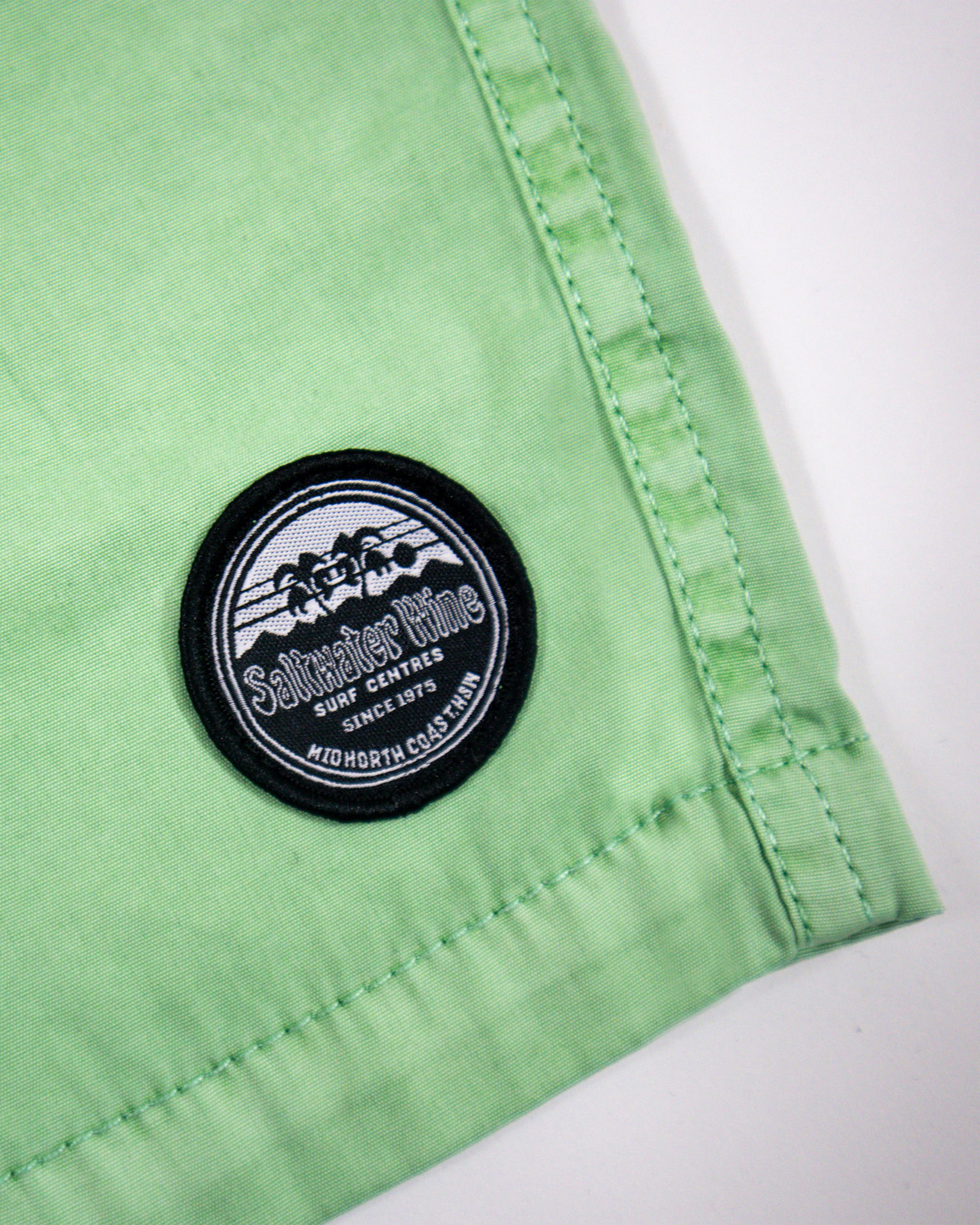 Close-up of Saltwater Wine logo patch on green board shorts hem.