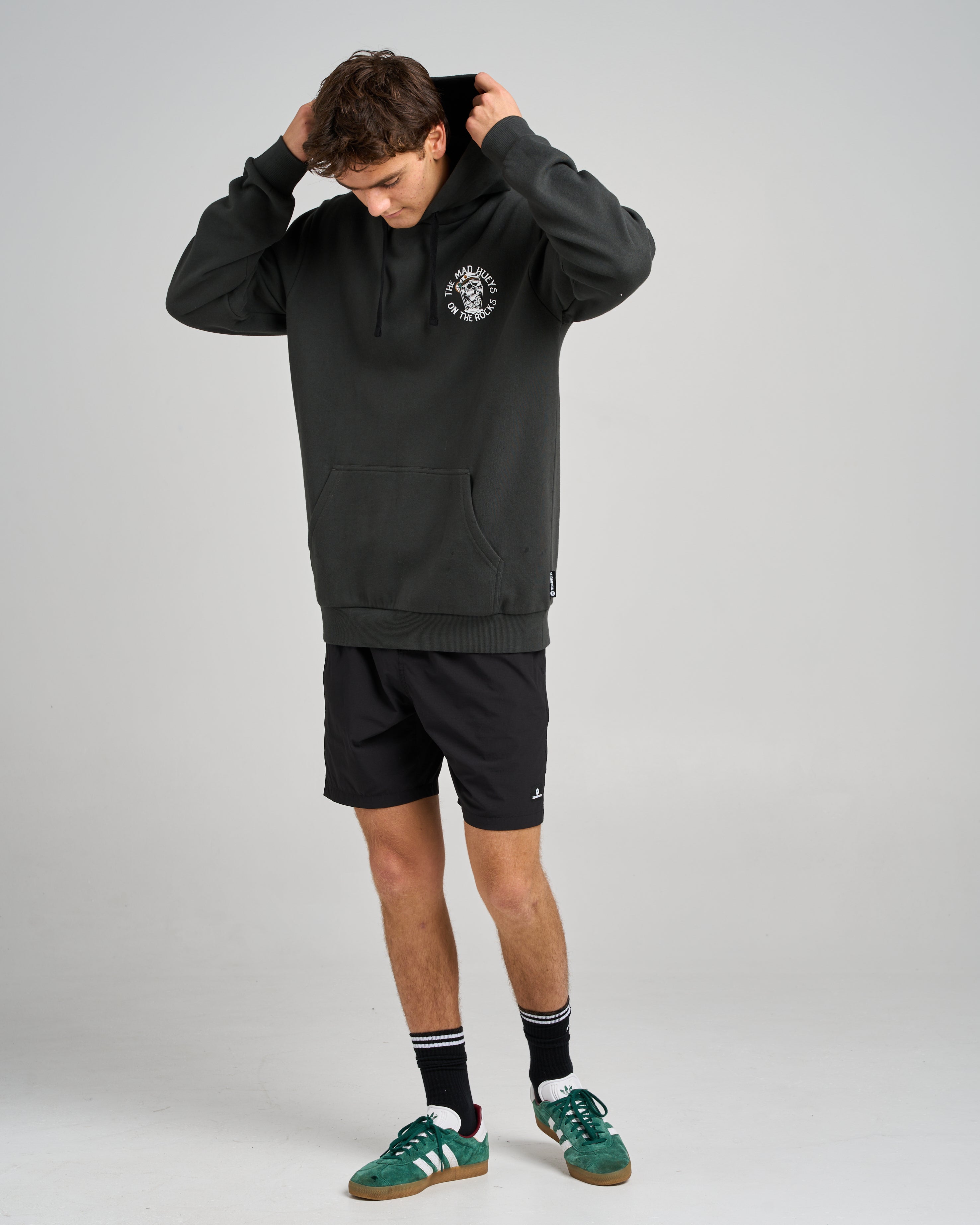Male model adjusting the hood of a black hoodie with a small white chest logo, paired with black shorts, socks, and green sneakers.