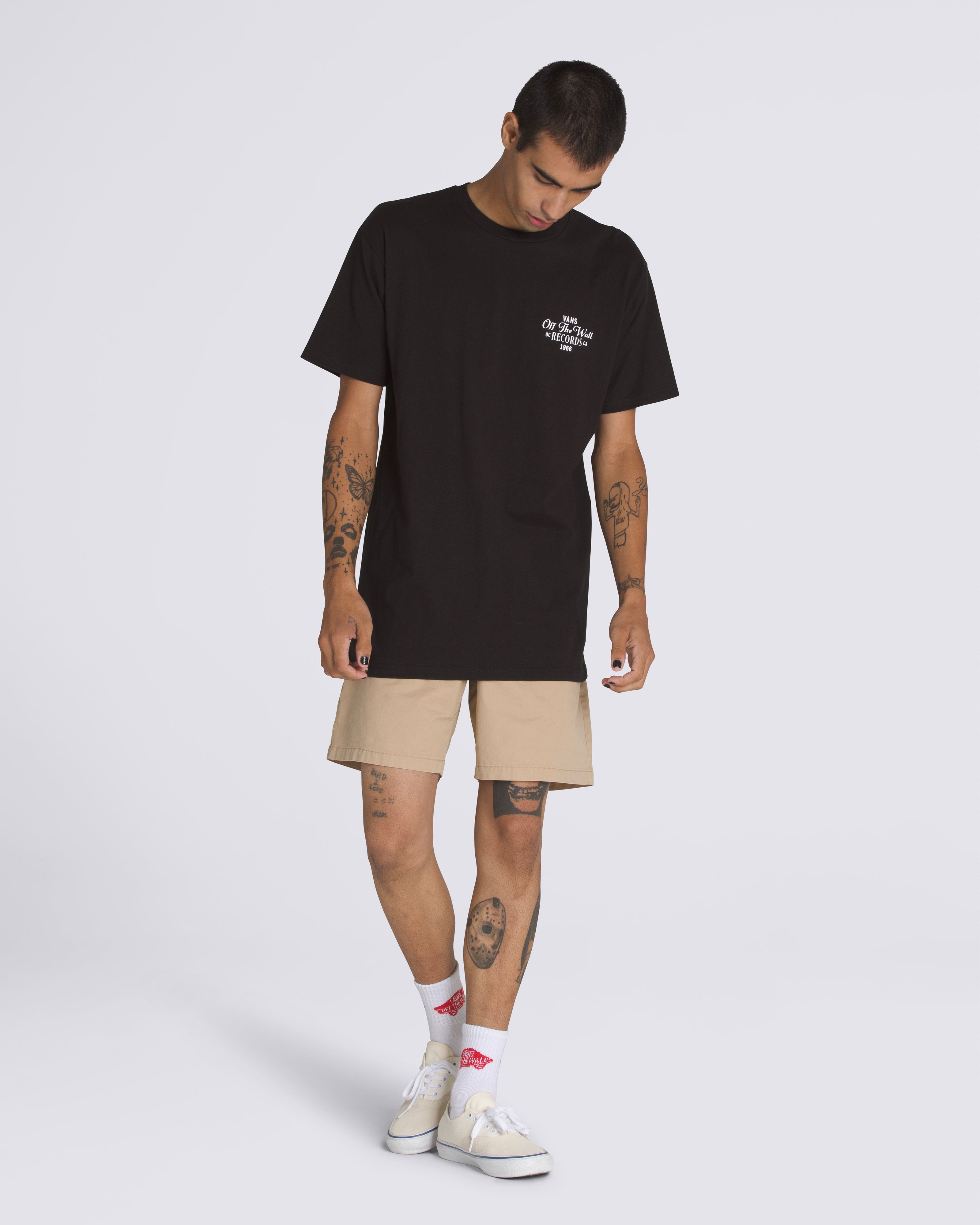 Model wearing a black Vans Off The Wall Records t-shirt with beige shorts, white socks, and white Vans sneakers.