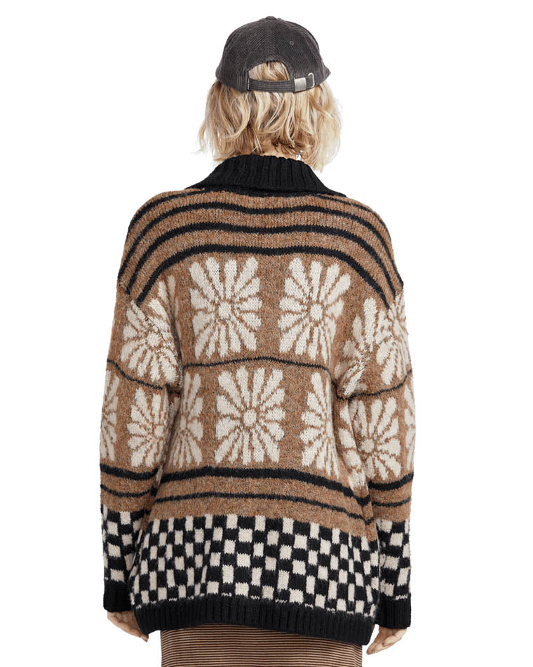 Back view of a knit cardigan featuring a floral and checkerboard pattern in earthy tones, worn by a model with a cap.