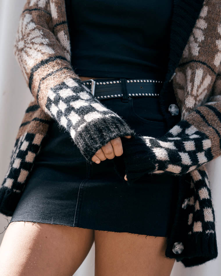 Close-up of a model wearing a knit cardigan with checkerboard cuffs, a black crop top, studded belt, and black denim skirt.
