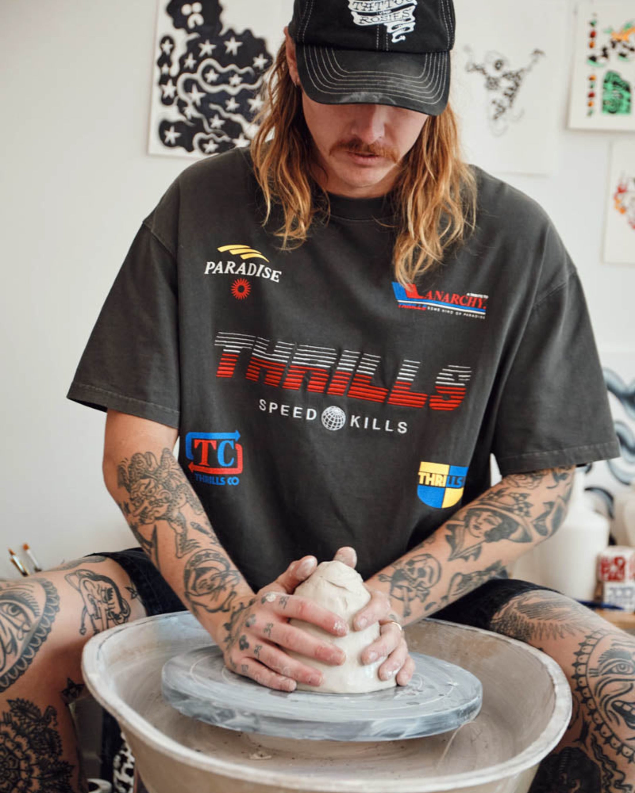 Person making a pot on a pottery wheel wearing a t-shirt with various logos.