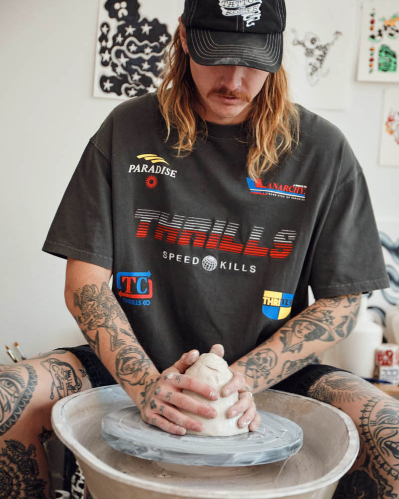 Person making a pot on a pottery wheel wearing a t-shirt with various logos.