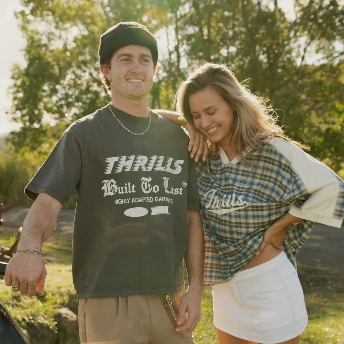 A man and a woman standing outdoors in a sunny park-like setting. The man wears a dark charcoal graphic T-shirt with white text reading 'THRILLS Built To Last' and khaki shorts, while the woman wears a blue plaid T-shirt over a white top with white shorts.