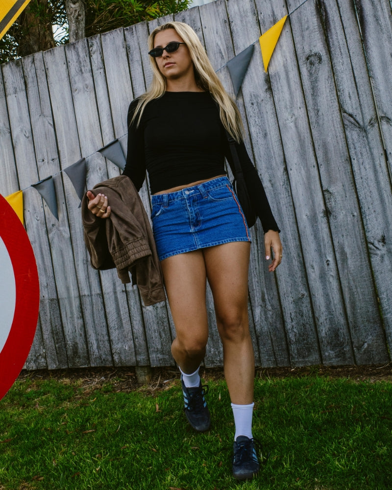 Woman in black crop top and blue denim skirt standing in front of a wooden fence with colorful flags.
