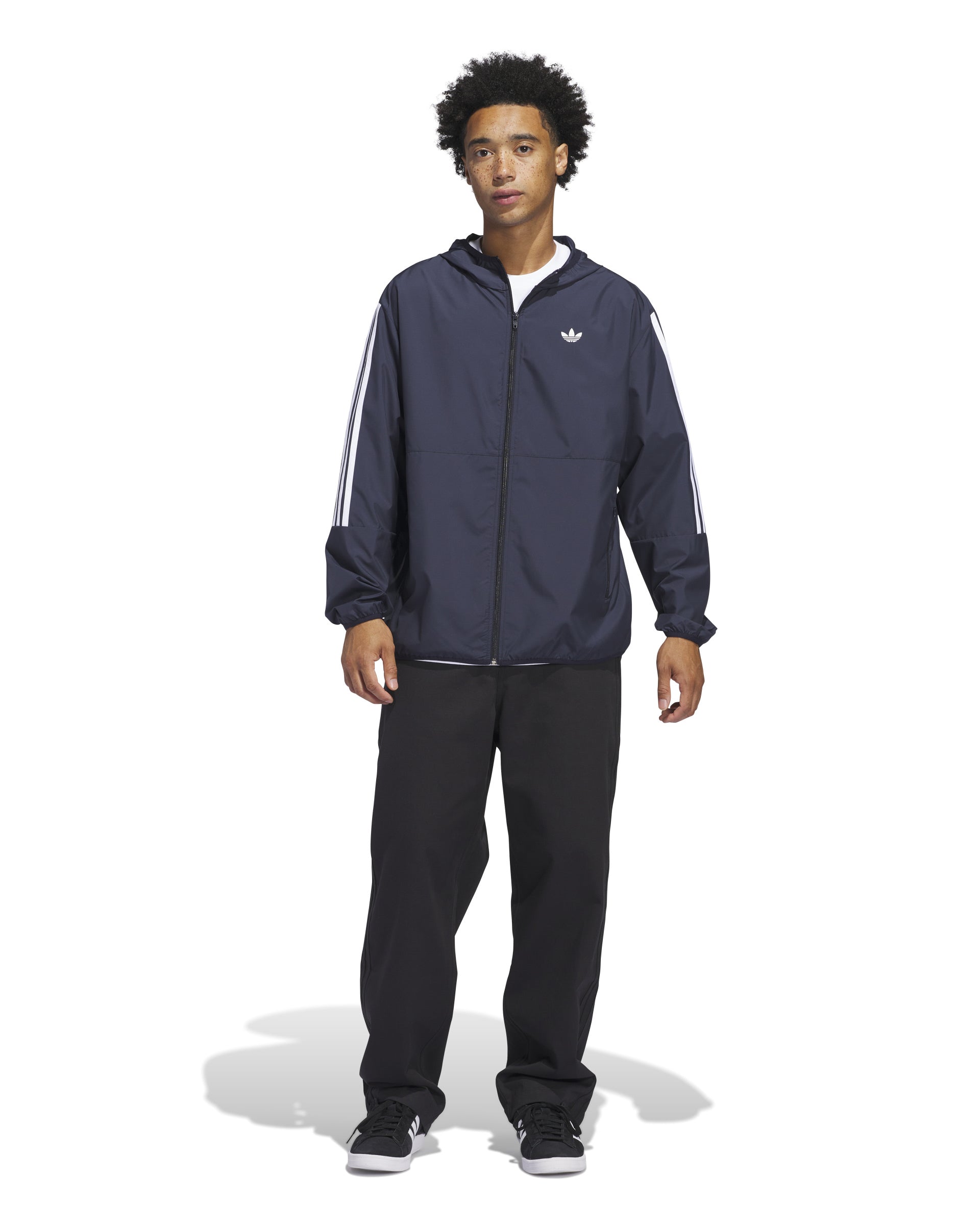 Full-body view of a man wearing a navy Adidas windbreaker jacket with white stripes on the sleeves, black pants, and sneakers.