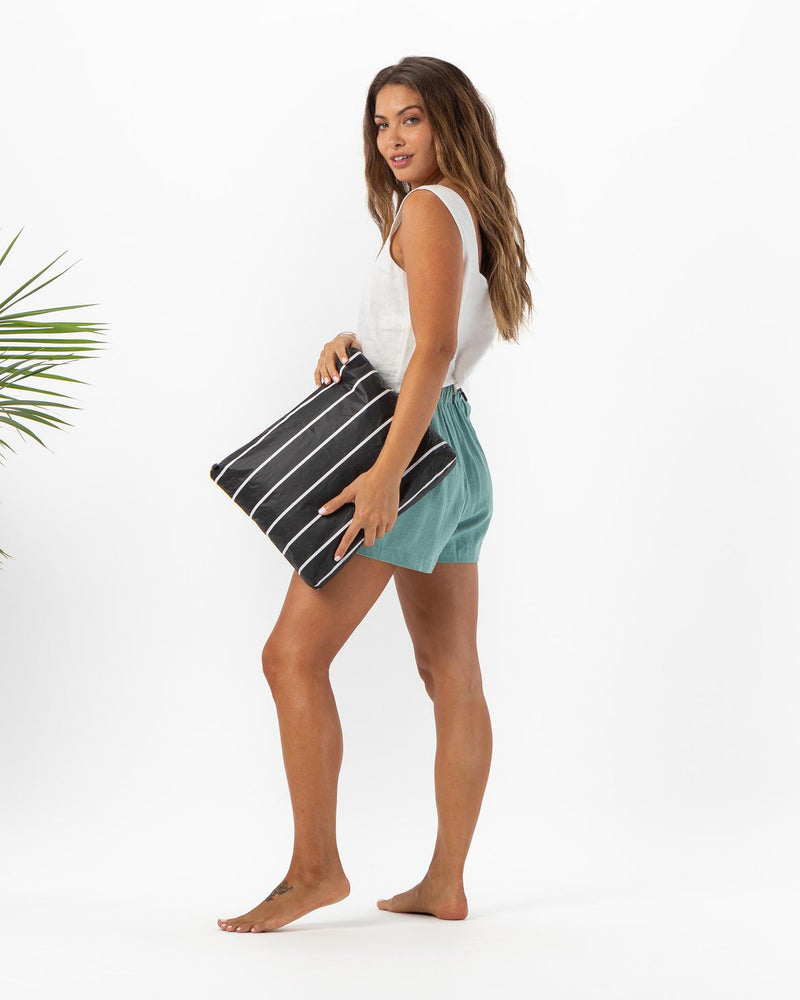 Woman carrying large black striped pouch while walking barefoot, styled in relaxed beachwear.