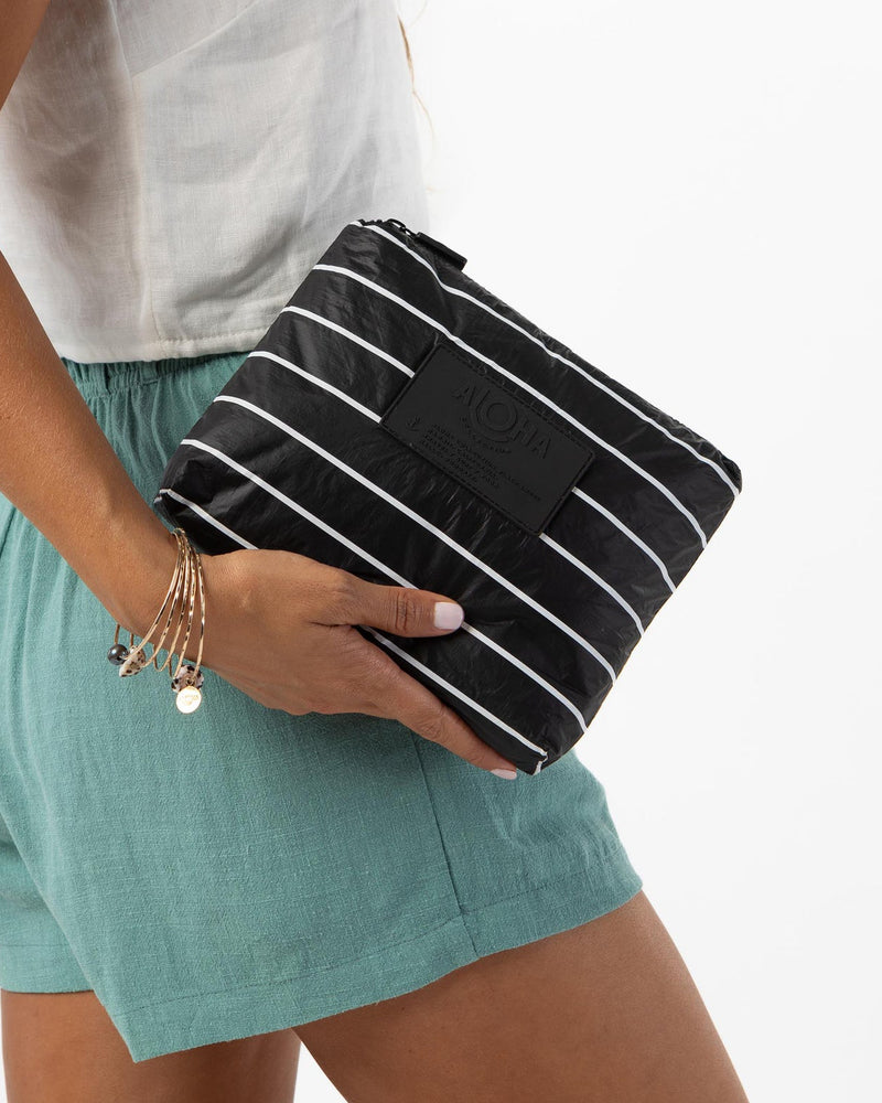 Woman holding black and white striped ALOHA pouch in one hand, wearing green shorts.