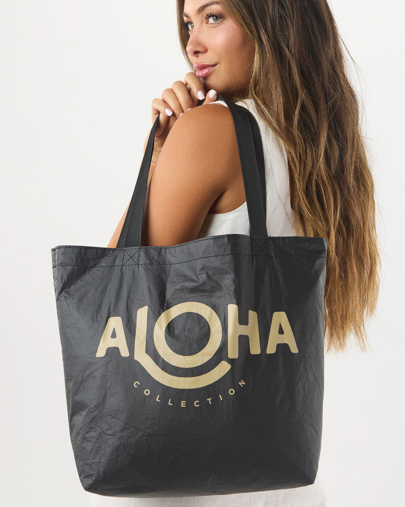 Woman holding reversible black tote bag with large beige ALOHA Collection logo