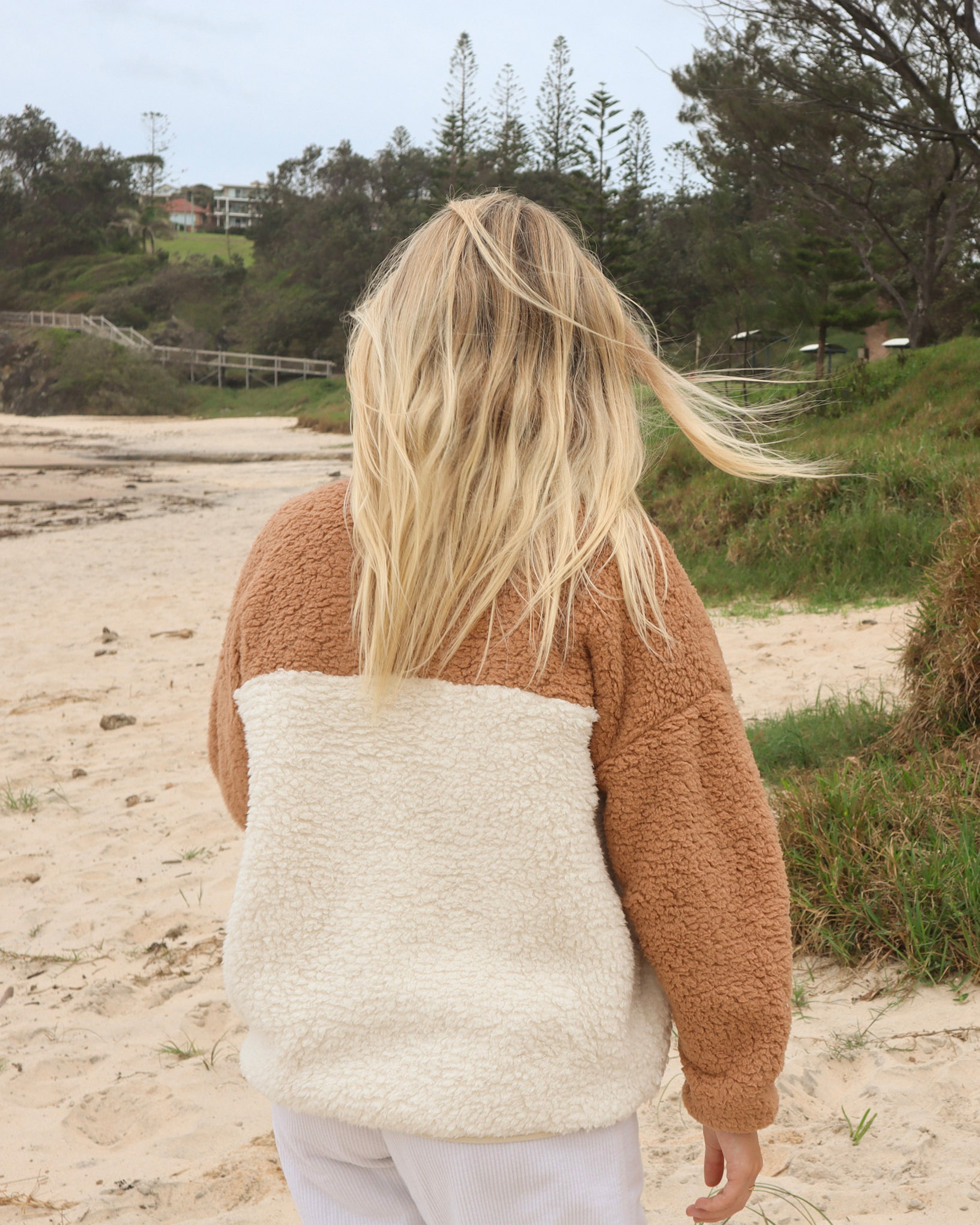 Back view of tan and cream sherpa fleece jacket worn on the beach, showcasing relaxed fit and cosy texture.
